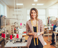 Foto: Eine Ausbilderin im Schneiderhandwerk in ihrem Nähatelier. Sie schaut lächelnd in die Kamera, hat ein Maßband um den Hals gelegt und ein Nadelkissen am Arm. 