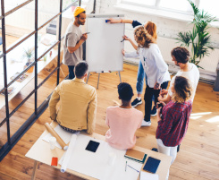 Foto: Eine Gruppe von Ausbilungspersonal oder Lehrkräften steht im Kreis um ein FlipChart und erarbeitet gemeinsam eine Strategie.