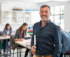 Foto: ein Lehrer an der Berufschule in seinem Klassenraum. Die Schüler/-innen im Hintergrund arbeiten an Aufgaben.