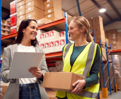 Foto: eine Ausbilderin und eine Auszubildende in einer Logistiklagerhalle sprechen miteinander. Die Ausbilderin hat ein Klemmbrett in der Hand, die Auszubildende trägt ein Paket.