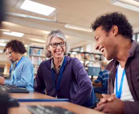 Foto: eine Lehrerin und ein Lehrer sitzen im Lehrerzimmer neben einander an Computern und lachen miteinander. Im Hintergrund ist das Lehrerzimmer mit Bücherregalen zu sehen.