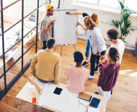 Foto: Eine Gruppe von Ausbilungspersonal oder Lehrkräften steht im Kreis um ein FlipChart und erarbeitet gemeinsam eine Strategie.